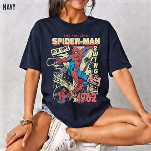 K&ouml;nnte beinhalten: Marineblaues T-Shirt mit einem Retro-Grafik von Spider-Man in New York City. Das Design enth&auml;lt den Text "The Amazing Spider-Man" und "1962". Das T-Shirt hat einen Vintage-Comic-Stil.