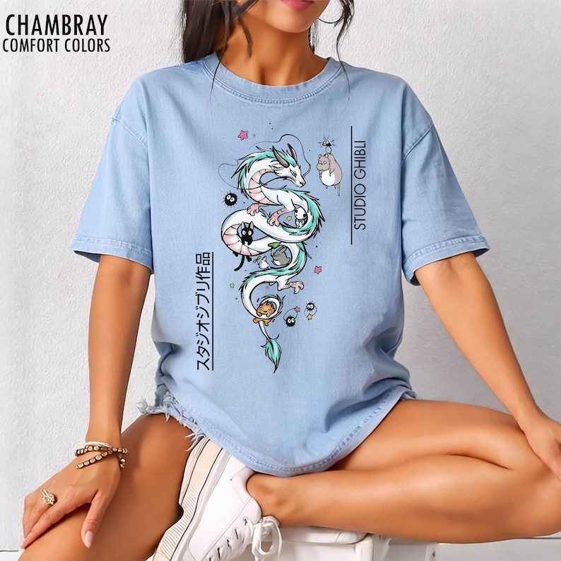May include: Light blue t-shirt with a Studio Ghibli design featuring a white dragon, black cats, and other characters. The shirt has the text "STUDIO GHIBLI" and Japanese text. The shirt is made of Chambray Comfort Colors.