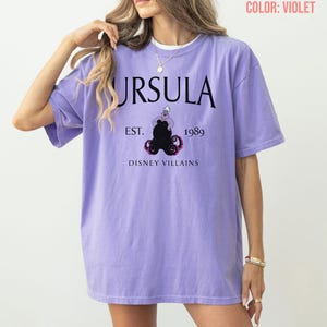 May include: A violet t-shirt with the word "URSULA" in black, and the text "EST. 1989" and "DISNEY VILLAINS" below. The graphic features a black Ursula illustration. The shirt has an oversized fit.