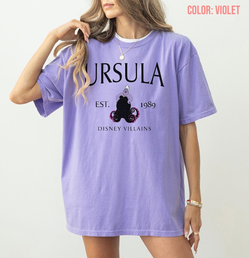 May include: A violet t-shirt with the word "URSULA" in black, and the text "EST. 1989" and "DISNEY VILLAINS" below. The graphic features a black Ursula illustration. The shirt has an oversized fit.