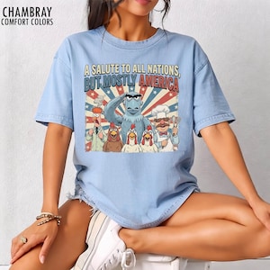 May include: Light blue t-shirt with the text "A Salute to All Nations, But Mostly America" and a graphic featuring cartoon characters. The graphic includes an eagle, chickens, and other characters in a patriotic design.
