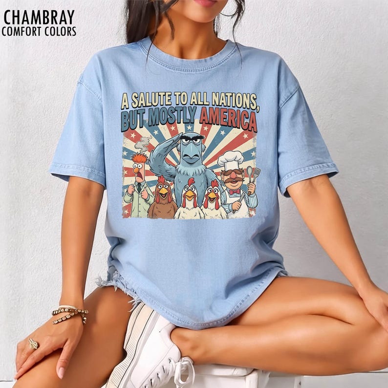 May include: Light blue t-shirt with the text "A Salute to All Nations, But Mostly America" and a graphic featuring cartoon characters. The graphic includes an eagle, chickens, and other characters in a patriotic design.