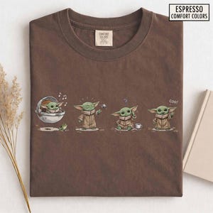 May include: Espresso-colored t-shirt featuring a cartoon depiction of a green alien character in various poses. The graphic includes the text "Espresso Comfort Colors" and the character is shown with a frog, a ball, and a cup.