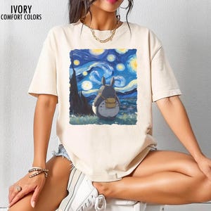 May include: Ivory-colored t-shirt with a graphic of a Totoro character looking at Van Gogh's "Starry Night" painting. The shirt has short sleeves and the words "IVORY COMFORT COLORS" printed at the top.