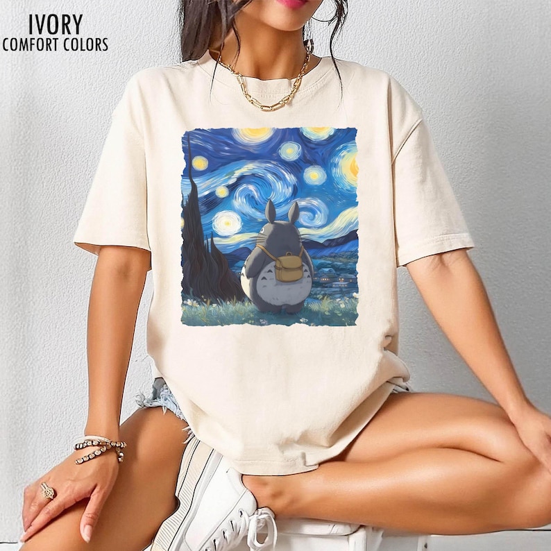 May include: Ivory-colored t-shirt with a graphic of a Totoro character looking at Van Gogh's "Starry Night" painting. The shirt has short sleeves and the words "IVORY COMFORT COLORS" printed at the top.
