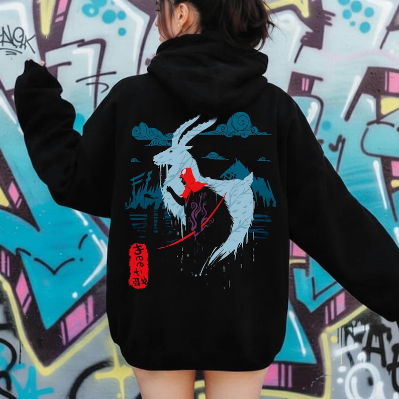 Peut inclure: Sweat &agrave; capuche noir avec une illustration stylis&eacute;e d'une cr&eacute;ature ressemblant &agrave; une ch&egrave;vre, v&ecirc;tue d'une cape rouge et d'&eacute;l&eacute;ments bleus. Le motif comprend du texte japonais et un paysage montagneux. Le sweat est port&eacute; par une personne.