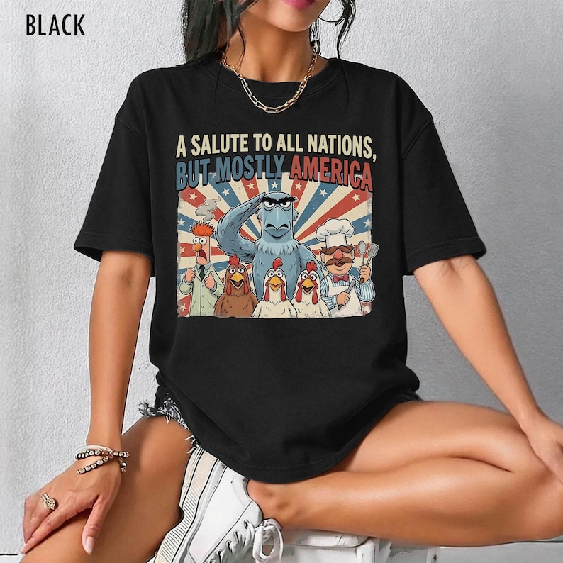 May include: Black t-shirt featuring a graphic with the text "A SALUTE TO ALL NATIONS, BUT MOSTLY AMERICA." The graphic includes cartoon characters of an eagle, chickens, and other figures in front of a red, white, and blue background.