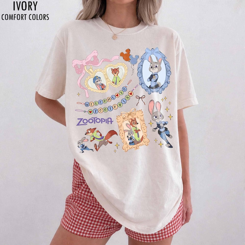 May include: An ivory short-sleeved t-shirt featuring a Zootopia graphic print. The design includes characters Judy Hopps and Nick Wilde, heart-shaped frames, a pink ribbon, and the text Welcome to Zootopia. The shirt has a crew neckline.