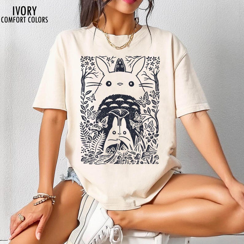 以下が含まれることがあります： アイボリー色のTシャツ。黒いグラフィックで、木、蝶、葉に囲まれたトトロのキャラクターが描かれています。「IVORY COMFORT COLORS」の文字が左上にプリントされています。半袖でゆったりとしたフィット感です。