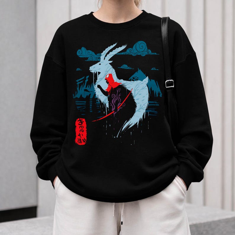 Peut inclure: Sweat-shirt noir avec un motif graphique d'une cr&eacute;ature ressemblant &agrave; une ch&egrave;vre bleu clair et une figure rouge, sur fond de nuages et de montagnes bleus. Texte japonais sur une &eacute;tiquette rouge. V&ecirc;tement d&eacute;contract&eacute;.
