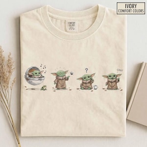 May include: Ivory Comfort Colors t-shirt featuring a four-panel illustration of a green alien character. The character is shown in a pod, playing with a ball, confused, and saying "coo!". The shirt has a crew neck and a tag that says "Comfort Colors".