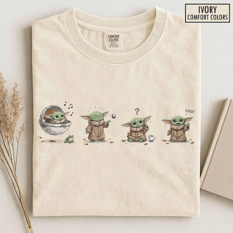 May include: Ivory Comfort Colors t-shirt featuring a four-panel illustration of a green alien character. The character is shown in a pod, playing with a ball, confused, and saying "coo!". The shirt has a crew neck and a tag that says "Comfort Colors".