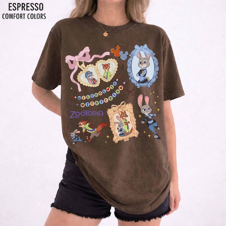 May include: An espresso brown short-sleeve t-shirt featuring a graphic print of Zootopia characters. The design includes framed portraits of Judy Hopps and Nick Wilde, a pink bow, and the text Welcome to Zootopia in colorful letters.