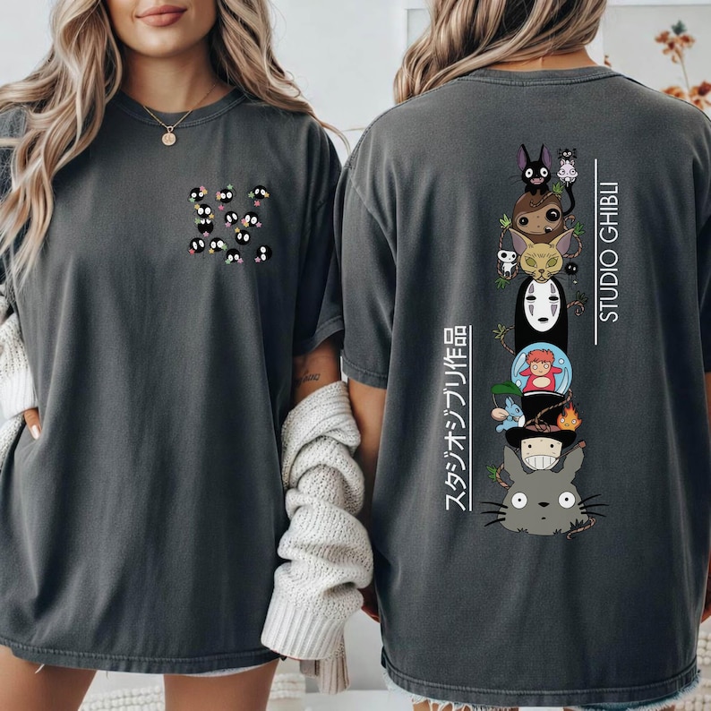 May include: Dark grey t-shirt featuring a small cluster of soot sprites on the front and a stacked graphic of Studio Ghibli characters on the back. The back graphic includes characters from various Ghibli films, with the text "STUDIO GHIBLI" and Japanese text.
