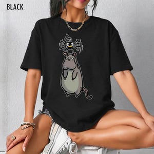May include: Black t-shirt with a cartoon design of a large, grey rat with a small bird on its head. The bird has spiky hair and is drawn in a whimsical style. The word "BLACK" is printed at the top of the shirt.