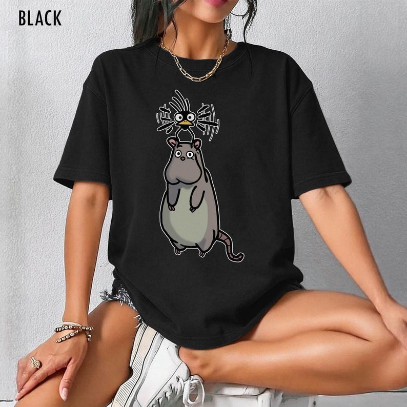 May include: Black t-shirt with a cartoon design of a large, grey rat with a small bird on its head. The bird has spiky hair and is drawn in a whimsical style. The word "BLACK" is printed at the top of the shirt.