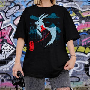 Peut inclure: T-shirt noir avec un motif d'une cr&eacute;ature blanche ressemblant &agrave; une ch&egrave;vre avec des accents rouges et du texte japonais. Le motif comprend des montagnes et des nuages bleus. Le t-shirt est port&eacute; par une personne.