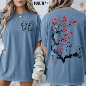 May include: Blue jean-colored t-shirt featuring a black graphic of three small figures on the front. The back of the shirt displays a tree with pink blossoms, figures, and Japanese text. The shirt has short sleeves and a relaxed fit.