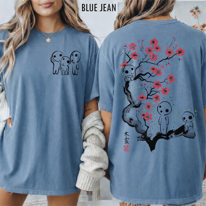May include: Blue jean-colored t-shirt featuring a black graphic of three small figures on the front. The back of the shirt displays a tree with pink blossoms, figures, and Japanese text. The shirt has short sleeves and a relaxed fit.
