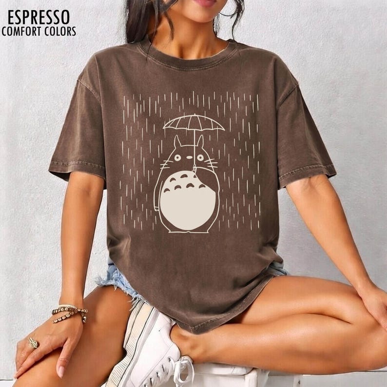 May include: Espresso-colored Comfort Colors t-shirt featuring a cartoon character holding an umbrella in the rain. The character is white with black details. The text "ESPRESSO COMFORT COLORS" is at the top left.