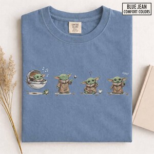 May include: Blue jean Comfort Colors t-shirt featuring a cartoon illustration of a character in various poses. The character is green with large ears and wears a brown robe. The shirt has the text "Blue Jean Comfort Colors".