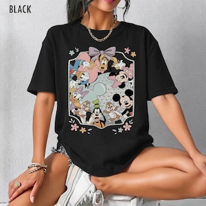 K&ouml;nnte beinhalten: Schwarzes T-Shirt mit einem farbenfrohen Aufdruck von Disney-Figuren wie Mickey Mouse, Minnie Mouse, Donald Duck, Goofy und Chip und Chap. Die Grafik wird von einem dekorativen Rahmen mit Blumen und einer Schleife umgeben.
