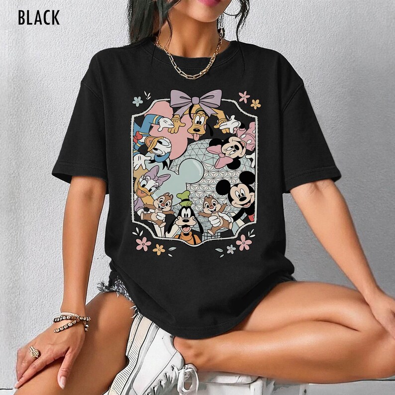 K&ouml;nnte beinhalten: Schwarzes T-Shirt mit einem farbenfrohen Aufdruck von Disney-Figuren wie Mickey Mouse, Minnie Mouse, Donald Duck, Goofy und Chip und Chap. Die Grafik wird von einem dekorativen Rahmen mit Blumen und einer Schleife umgeben.