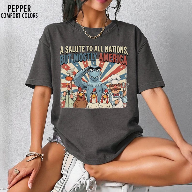 May include: Dark gray t-shirt featuring a graphic of Muppet characters, including Sam Eagle, chickens, and the Swedish Chef. The text reads "A Salute to All Nations, But Mostly America" in a patriotic design.