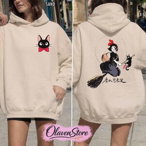 May include: Sand-colored hoodie featuring a black cat with a red bow tie on the front. The back displays a graphic of a girl on a broom with a black cat and Japanese text. The hoodie has a kangaroo pocket and a hood.
