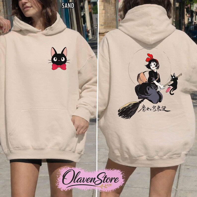 May include: Sand-colored hoodie featuring a black cat with a red bow tie on the front. The back displays a graphic of a girl on a broom with a black cat and Japanese text. The hoodie has a kangaroo pocket and a hood.