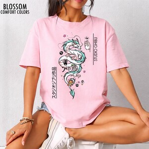 May include: A light pink t-shirt featuring a Studio Ghibli design. The graphic includes a white dragon with teal accents, and other characters. The text "STUDIO GHIBLI" and Japanese text are printed vertically. The shirt has the text "BLOSSOM COMFORT COLORS" at the top left.