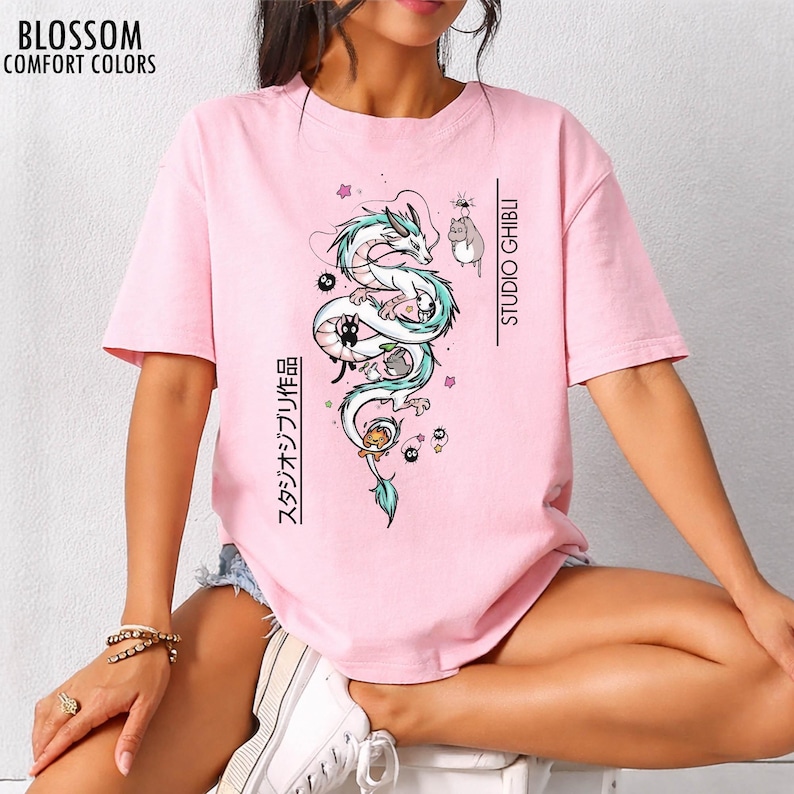 May include: A light pink t-shirt featuring a Studio Ghibli design. The graphic includes a white dragon with teal accents, and other characters. The text "STUDIO GHIBLI" and Japanese text are printed vertically. The shirt has the text "BLOSSOM COMFORT COLORS" at the top left.