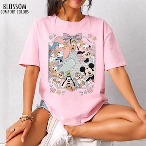 K&ouml;nnte beinhalten: Hellrosa T-Shirt mit einem farbenfrohen Aufdruck von Disney-Figuren, darunter Micky Maus, Goofy und Donald Duck. Die Grafik ist von einem dekorativen Rahmen mit Blumen und einer Schleife umgeben. Der Text "BLOSSOM COMFORT COLORS" ist oben links aufgedruckt.