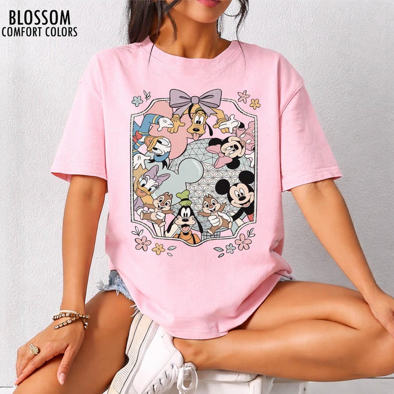 K&ouml;nnte beinhalten: Hellrosa T-Shirt mit einem farbenfrohen Aufdruck von Disney-Figuren, darunter Micky Maus, Goofy und Donald Duck. Die Grafik ist von einem dekorativen Rahmen mit Blumen und einer Schleife umgeben. Der Text "BLOSSOM COMFORT COLORS" ist oben links aufgedruckt.