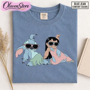 Op de afbeelding: Blauw Comfort Colors T-shirt met een cartoon illustratie van Stitch en Lilo met een zonnebril. Het shirt heeft de tekst "BLUE JEAN COMFORT COLORS" in de rechterbovenhoek. Het OlavenStore-logo staat in de linkerbovenhoek.