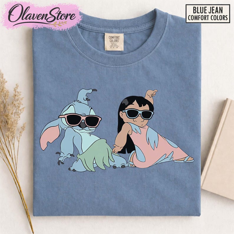Op de afbeelding: Blauw Comfort Colors T-shirt met een cartoon illustratie van Stitch en Lilo met een zonnebril. Het shirt heeft de tekst "BLUE JEAN COMFORT COLORS" in de rechterbovenhoek. Het OlavenStore-logo staat in de linkerbovenhoek.