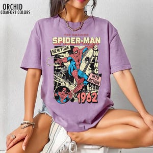 K&ouml;nnte beinhalten: Ein lila T-Shirt mit einem Vintage-Comic-Grafik von Spider-Man. Das Design enth&auml;lt den Text "THE AMAZING SPIDER-MAN", "NEW YORK" und "1962". Der Superheld ist in seinem klassischen rot-blauen Anzug dargestellt.
