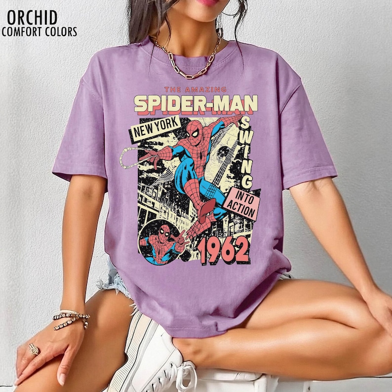 K&ouml;nnte beinhalten: Ein lila T-Shirt mit einem Vintage-Comic-Grafik von Spider-Man. Das Design enth&auml;lt den Text "THE AMAZING SPIDER-MAN", "NEW YORK" und "1962". Der Superheld ist in seinem klassischen rot-blauen Anzug dargestellt.