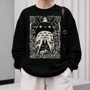以下が含まれることがあります： 黒いスウェットシャツ。クリーム色のグラフィックで、アニメ映画「となりのトトロ」のキャラクターが登場する森のシーンを描いています。デザインにはトトロ、蝶、葉が含まれています。
