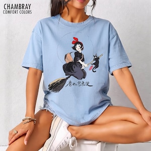 May include: Light blue t-shirt featuring a cartoon illustration of a young witch riding a broom with a black cat. The witch wears a black dress and a red bow. Japanese text is below the illustration. The shirt has the text "CHAMBRAY COMFORT COLORS" at the top.