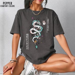 May include: Dark gray t-shirt featuring a white and teal dragon illustration with black cats and other characters. The text "STUDIO GHIBLI" and Japanese text are printed on the shirt. The shirt is from Pepper Comfort Colors.