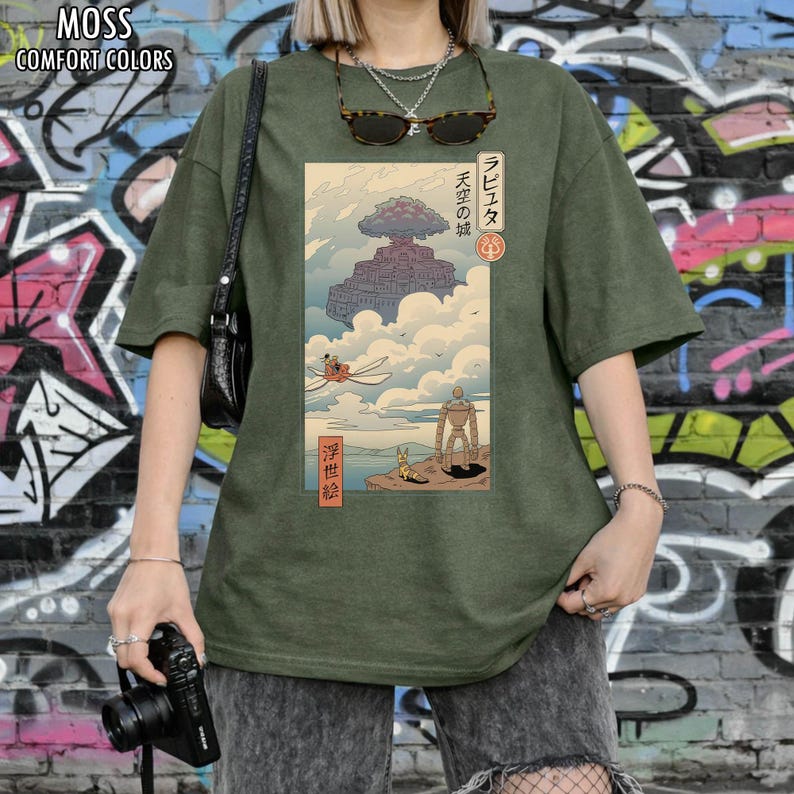 May include: A moss green Comfort Colors t-shirt with a Japanese anime-style illustration. The graphic shows a floating castle, a flying machine, and a robot. The text on the shirt includes Japanese characters and the words "Comfort Colors".