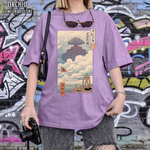 May include: A lilac-coloured t-shirt from Orchid Comfort Colors, with a Japanese anime-style illustration. The graphic shows a floating island with characters and Japanese text. The tee has short sleeves and a relaxed fit.