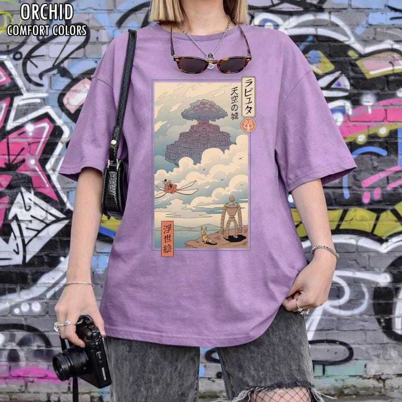 May include: A lilac-coloured t-shirt from Orchid Comfort Colors, with a Japanese anime-style illustration. The graphic shows a floating island with characters and Japanese text. The tee has short sleeves and a relaxed fit.