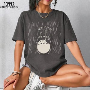 May include: Dark grey t-shirt featuring a white illustration of a Totoro character holding an umbrella in the rain. The text "PEPPER COMFORT COLORS" is printed at the top left. The shirt has short sleeves and a relaxed fit.