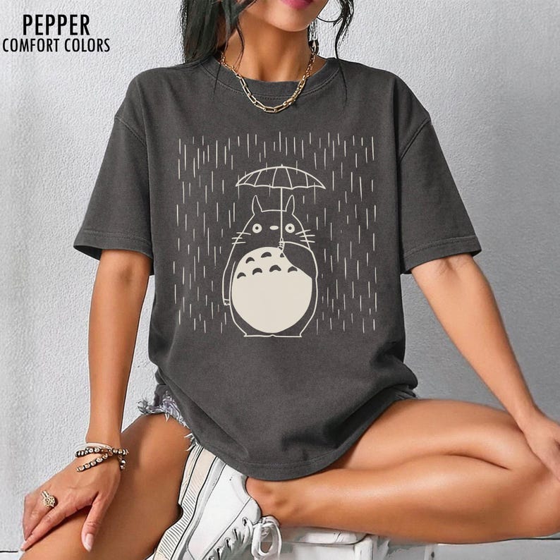 May include: Dark grey t-shirt featuring a white illustration of a Totoro character holding an umbrella in the rain. The text "PEPPER COMFORT COLORS" is printed at the top left. The shirt has short sleeves and a relaxed fit.