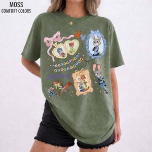May include: A moss green short-sleeve t-shirt featuring a graphic print of Zootopia characters. The design includes framed portraits of Judy Hopps and Nick Wilde, a pink ribbon, and the text Welcome to Zootopia in colorful letters.