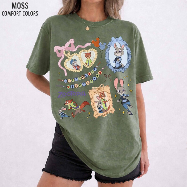May include: A moss green short-sleeve t-shirt featuring a graphic print of Zootopia characters. The design includes framed portraits of Judy Hopps and Nick Wilde, a pink ribbon, and the text Welcome to Zootopia in colorful letters.