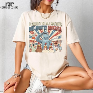 May include: Ivory-colored t-shirt with the text "A Salute to All Nations, But Mostly America." The graphic features cartoon characters including an eagle, chickens, and a chef. The shirt also has the words "Ivory Comfort Colors" printed on it.