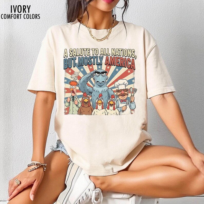 May include: Ivory-colored t-shirt with the text "A Salute to All Nations, But Mostly America." The graphic features cartoon characters including an eagle, chickens, and a chef. The shirt also has the words "Ivory Comfort Colors" printed on it.